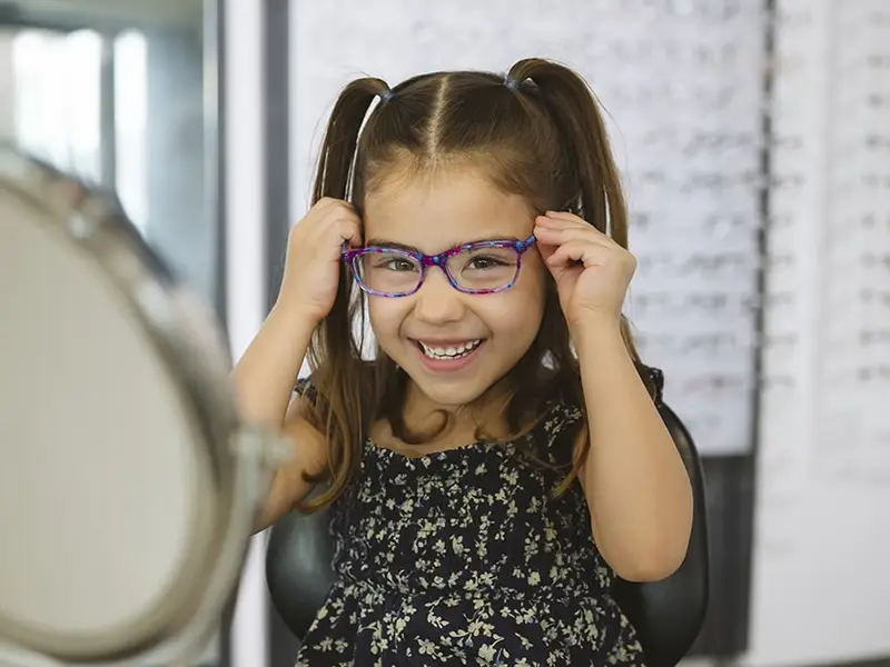 A young girl smiling as she tries on a pair of patterned glasses in front of a mirror, with frame displays behind her.